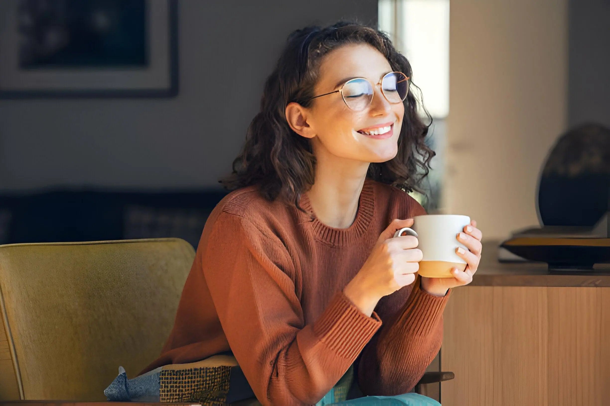 Mulher sorridente com óculos segurando uma xícara de chá em ambiente aconchegante.