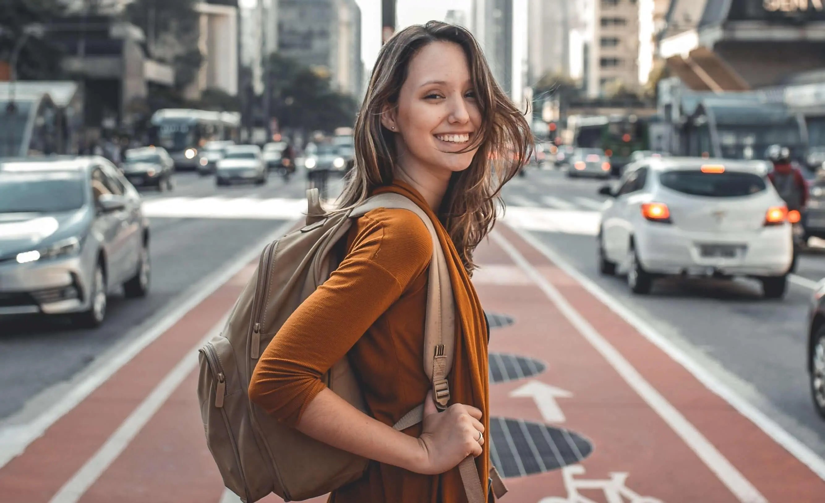 Texto alternativo:Mulher sorridente com mochila em avenida movimentada, simbolizando qualidade de vida urbana.