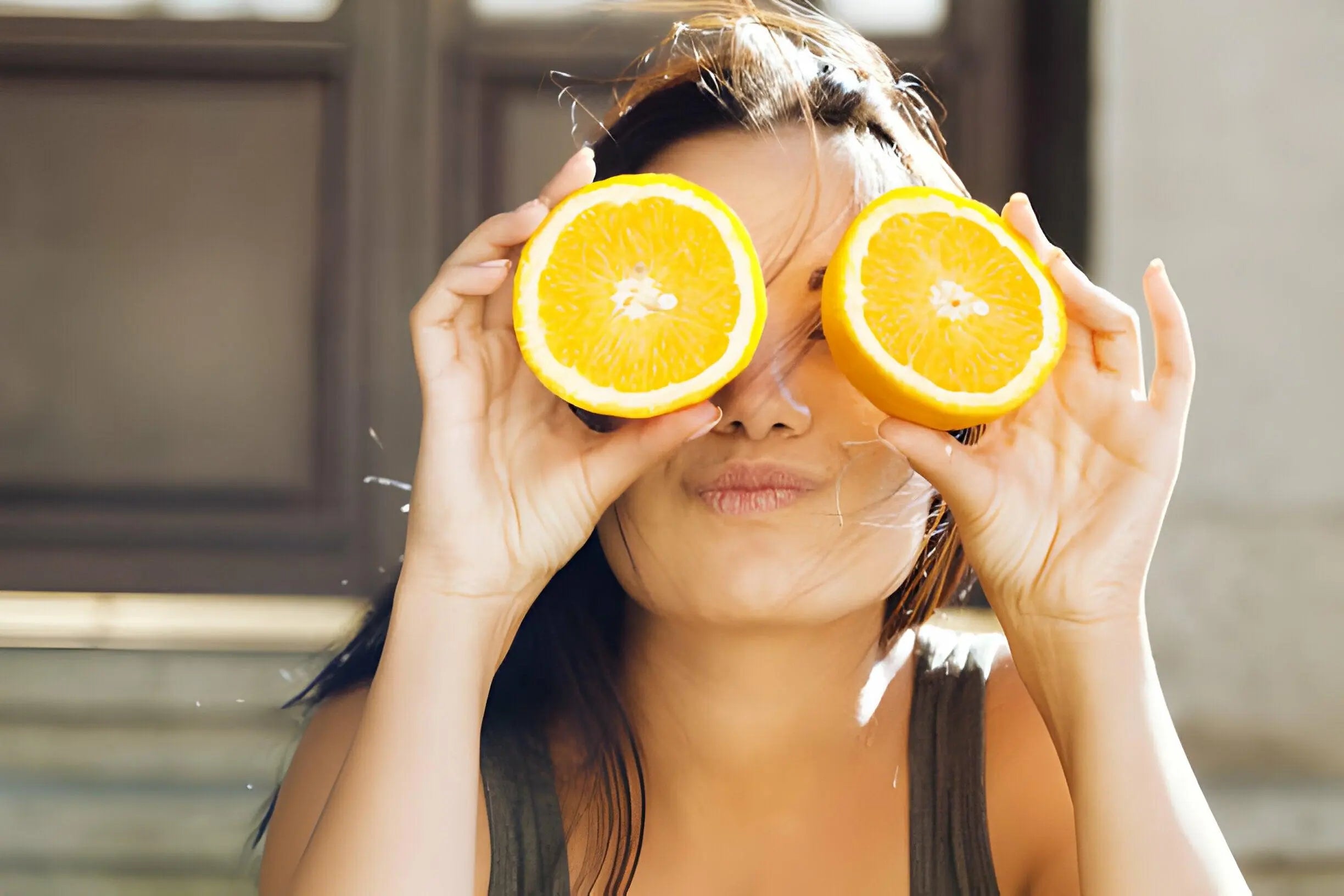 Texto alternativo:Mulher sorridente segurando metades de laranja nos olhos, simbolizando reposição de vitaminas no verão.