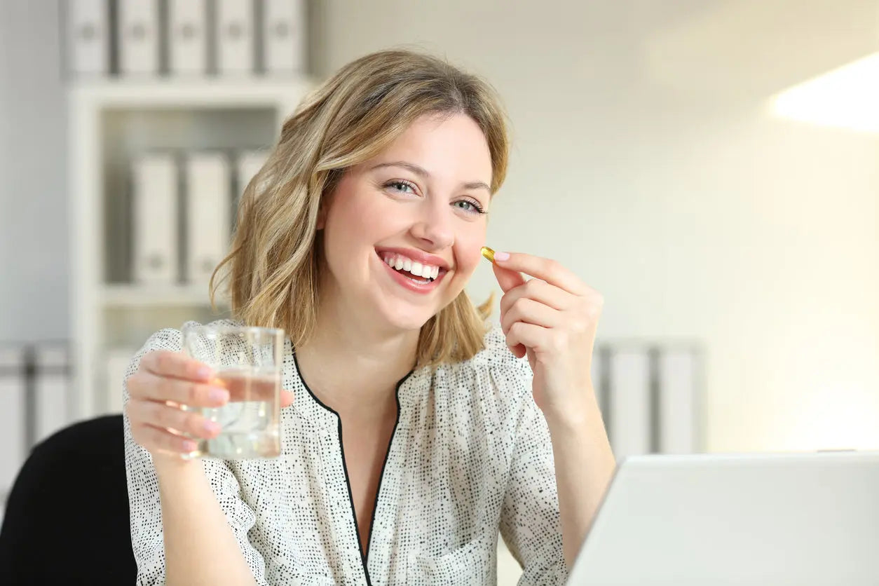 Mulher sorridente segurando cápsula de suplemento e copo d'água, simbolizando saúde.