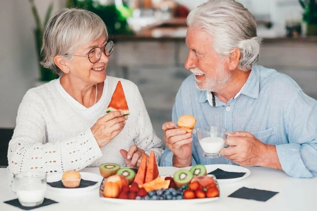 Casal de idosos sorrindo enquanto compartilham frutas e alimentos saudáveis.