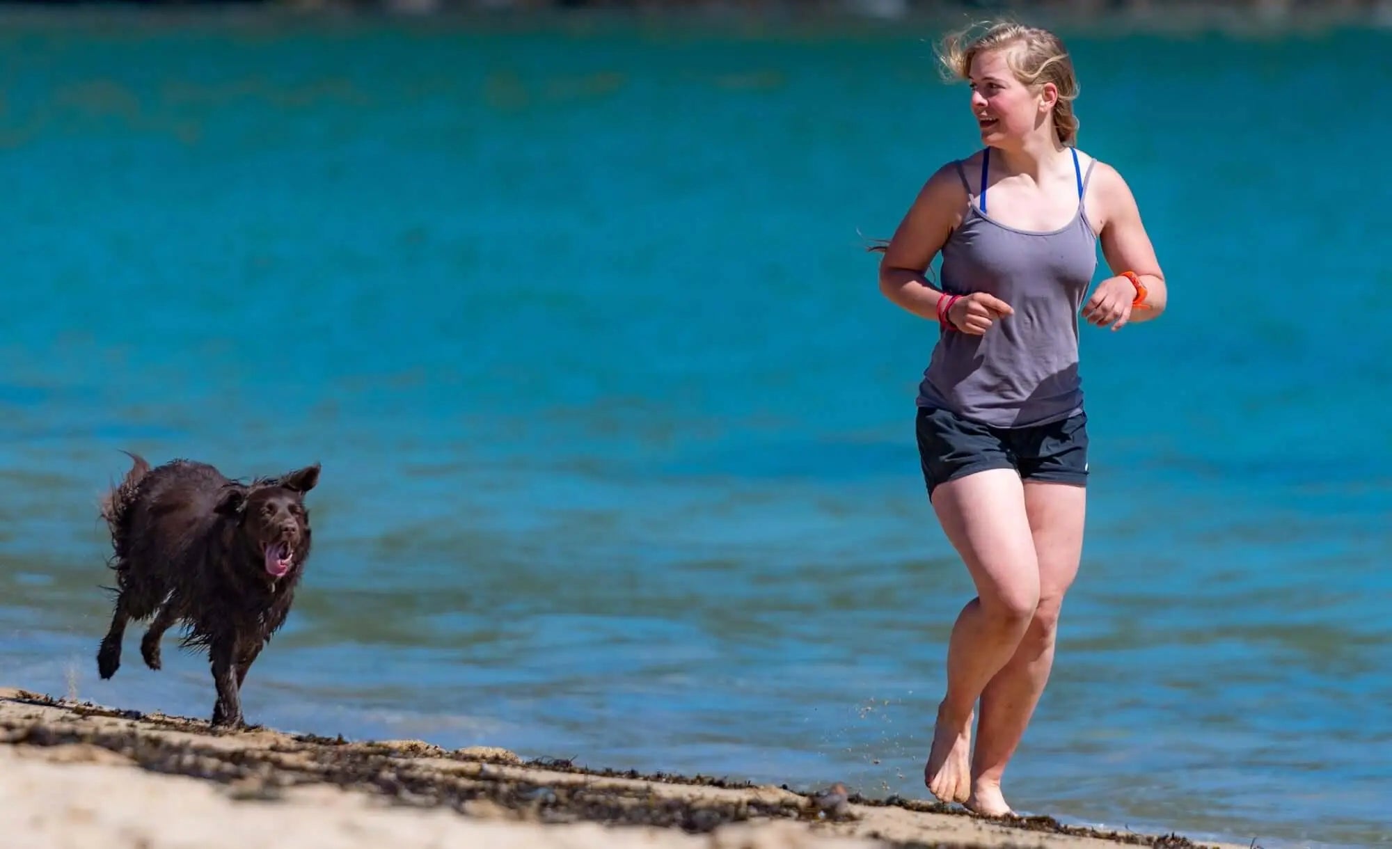 Mulher correndo descalça na praia ao lado de um cachorro marrom, com mar azul ao fundo.