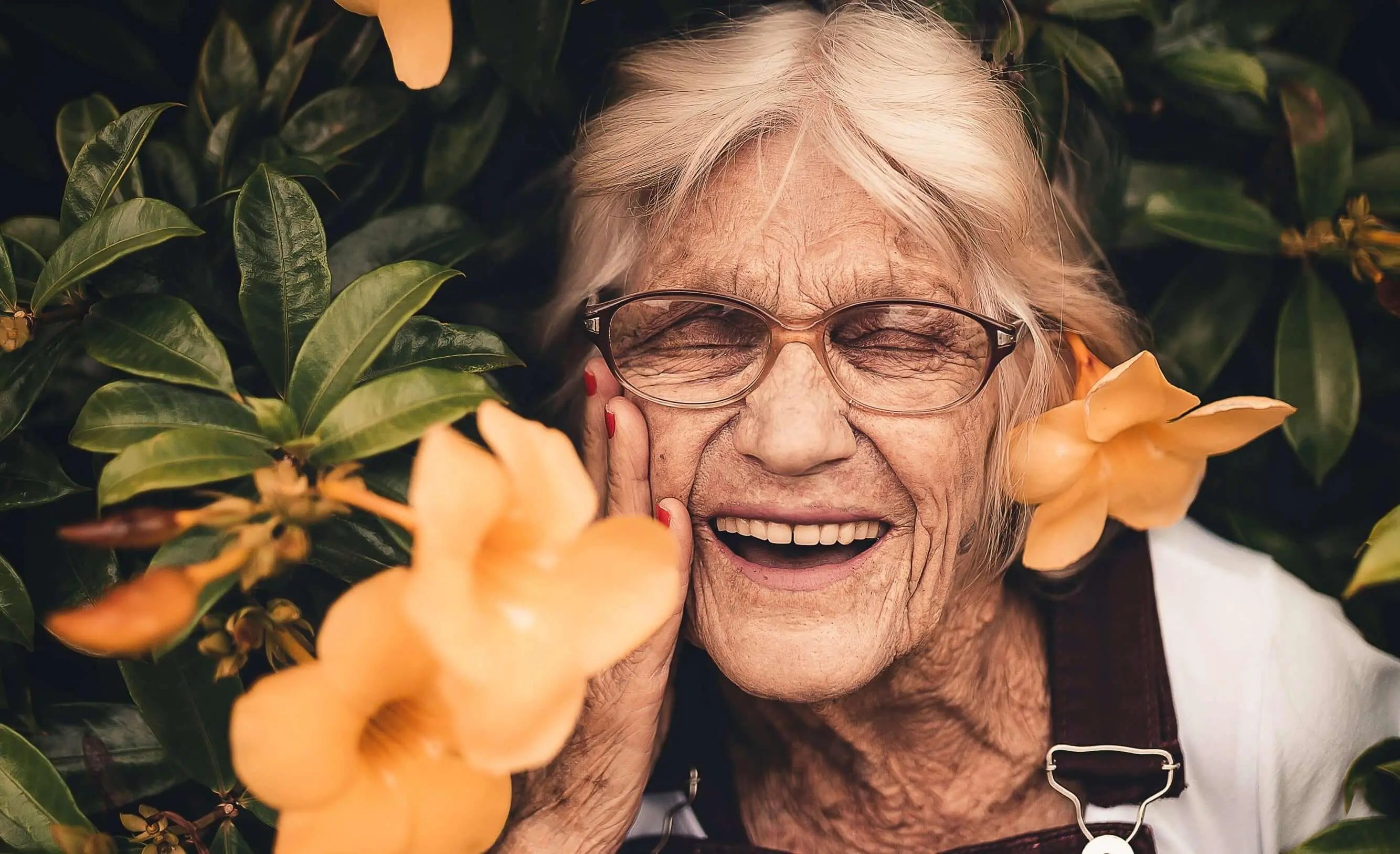 Mulher sorridente com óculos entre flores, representando vitalidade após os 50 anos.
