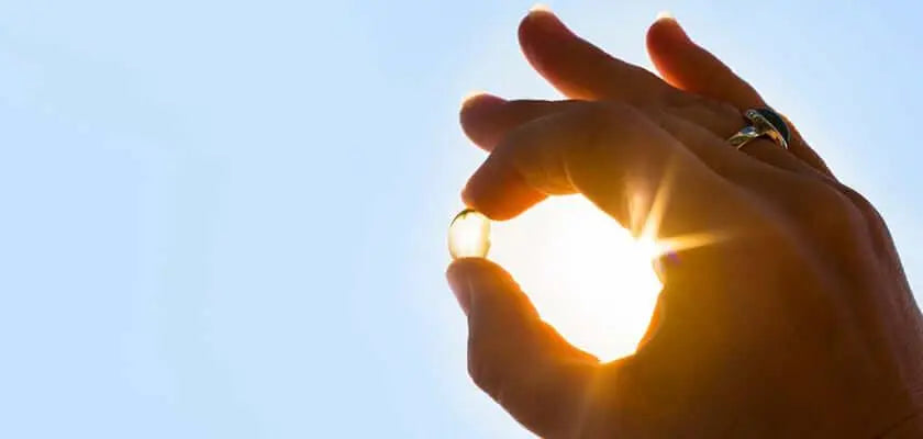 Texto alternativo:Mão segurando cápsula de vitamina D contra o sol, destacando sua importância.