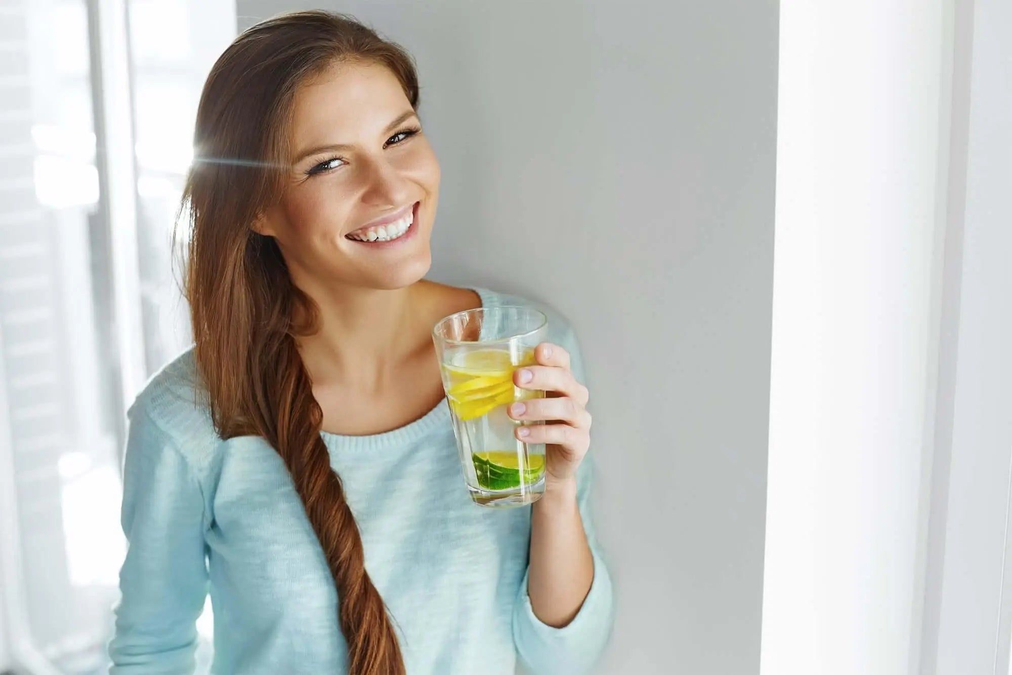 Texto alternativo:Mulher sorridente segurando um copo de água com limão, simbolizando saúde e bem-estar.