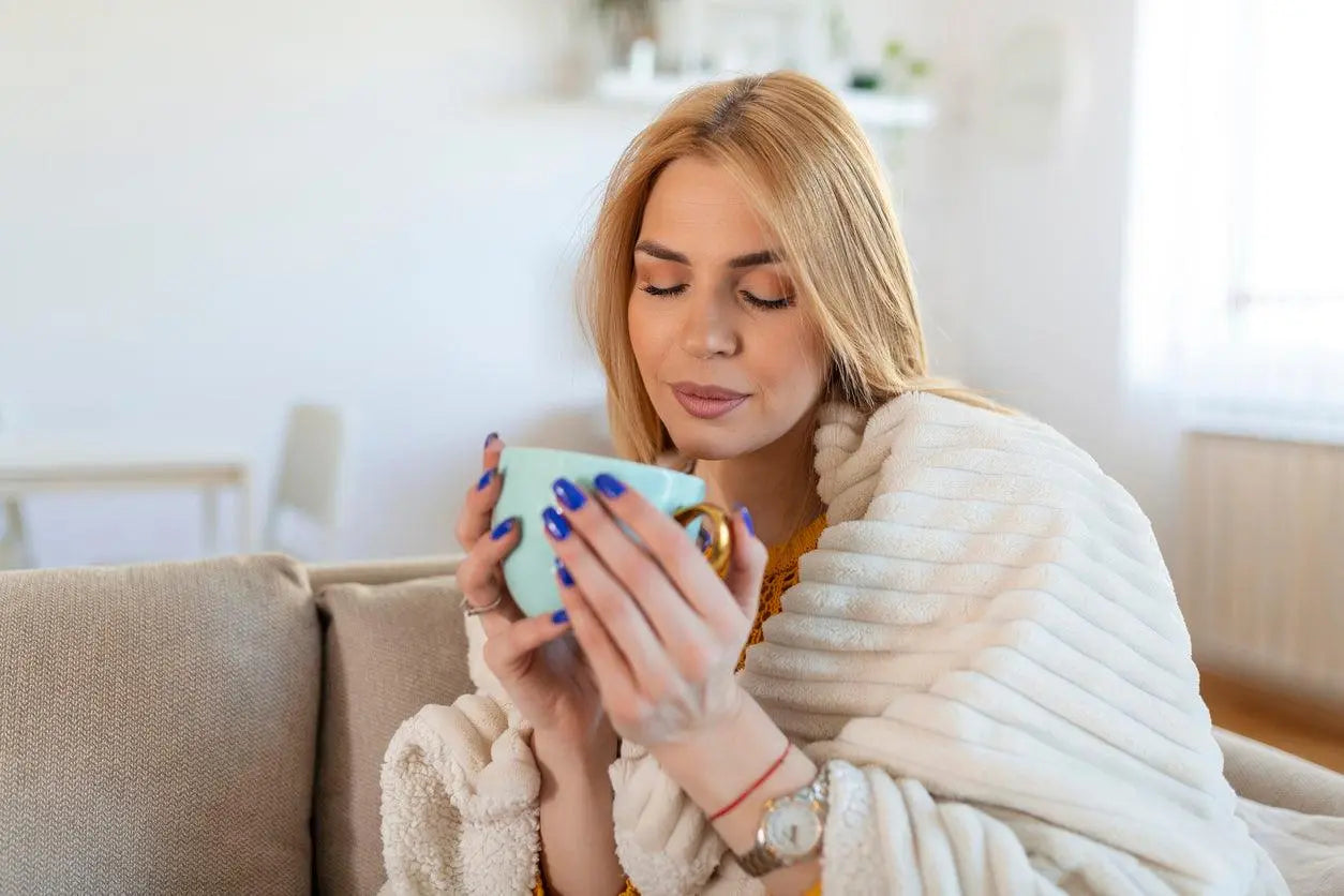 Texto alternativo:Mulher relaxando no inverno com xícara de chá, envolta em cobertor.