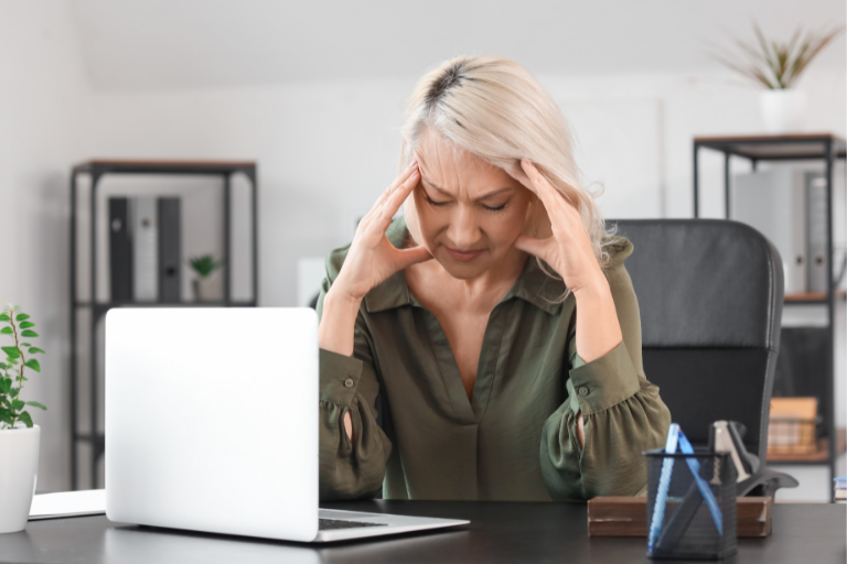 Mulher madura com expressão de desconforto, mãos na cabeça, sentada em frente ao computador.