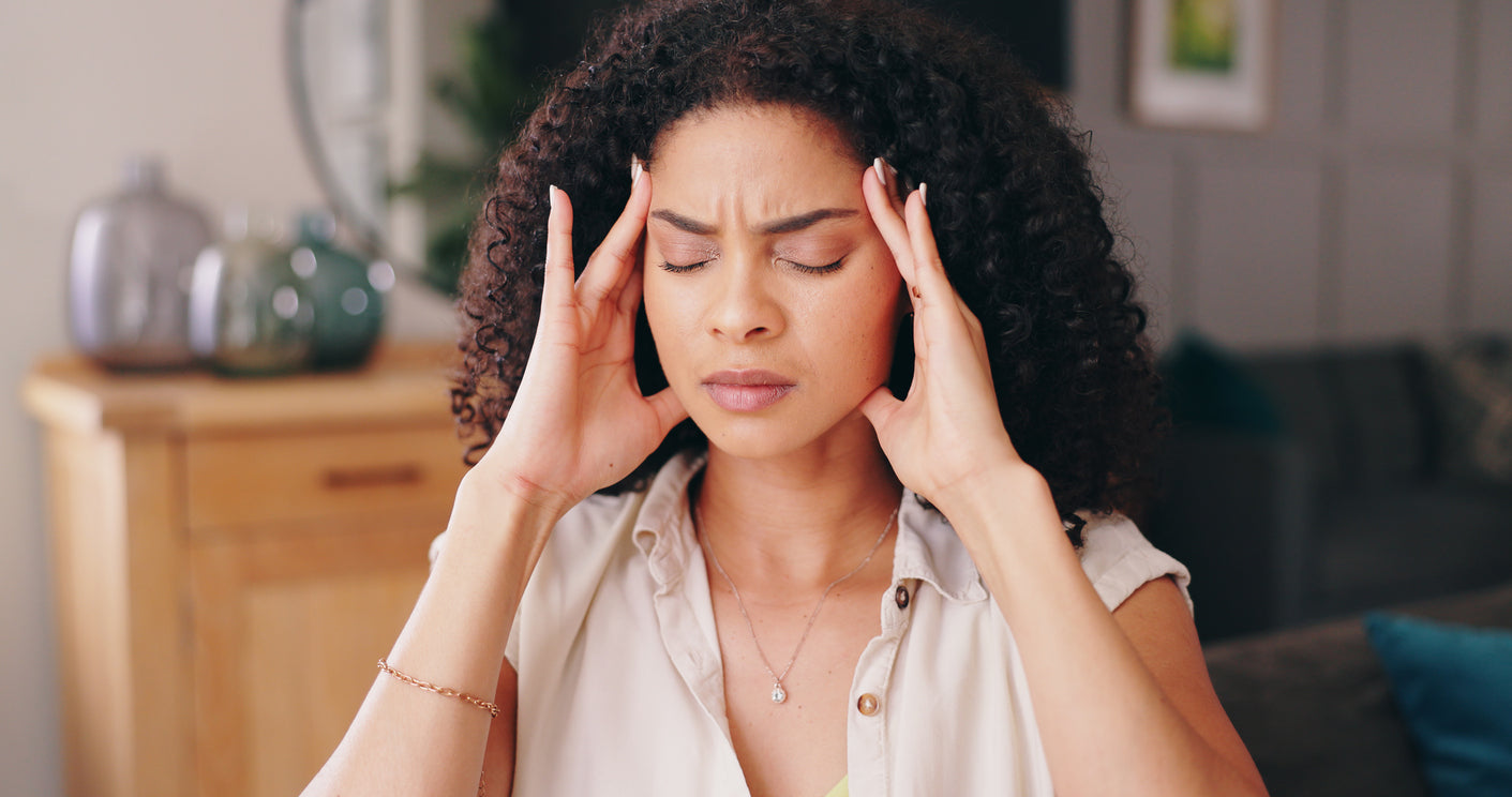 Mulher com expressão de dor de cabeça, tocando as têmporas, em ambiente doméstico.