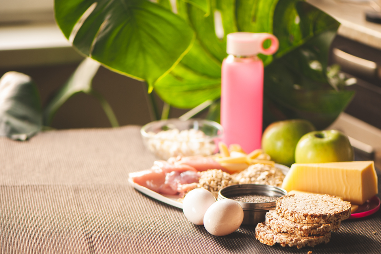Alimentos saudáveis como ovos, grãos, frutas e queijo, com garrafa rosa ao fundo.