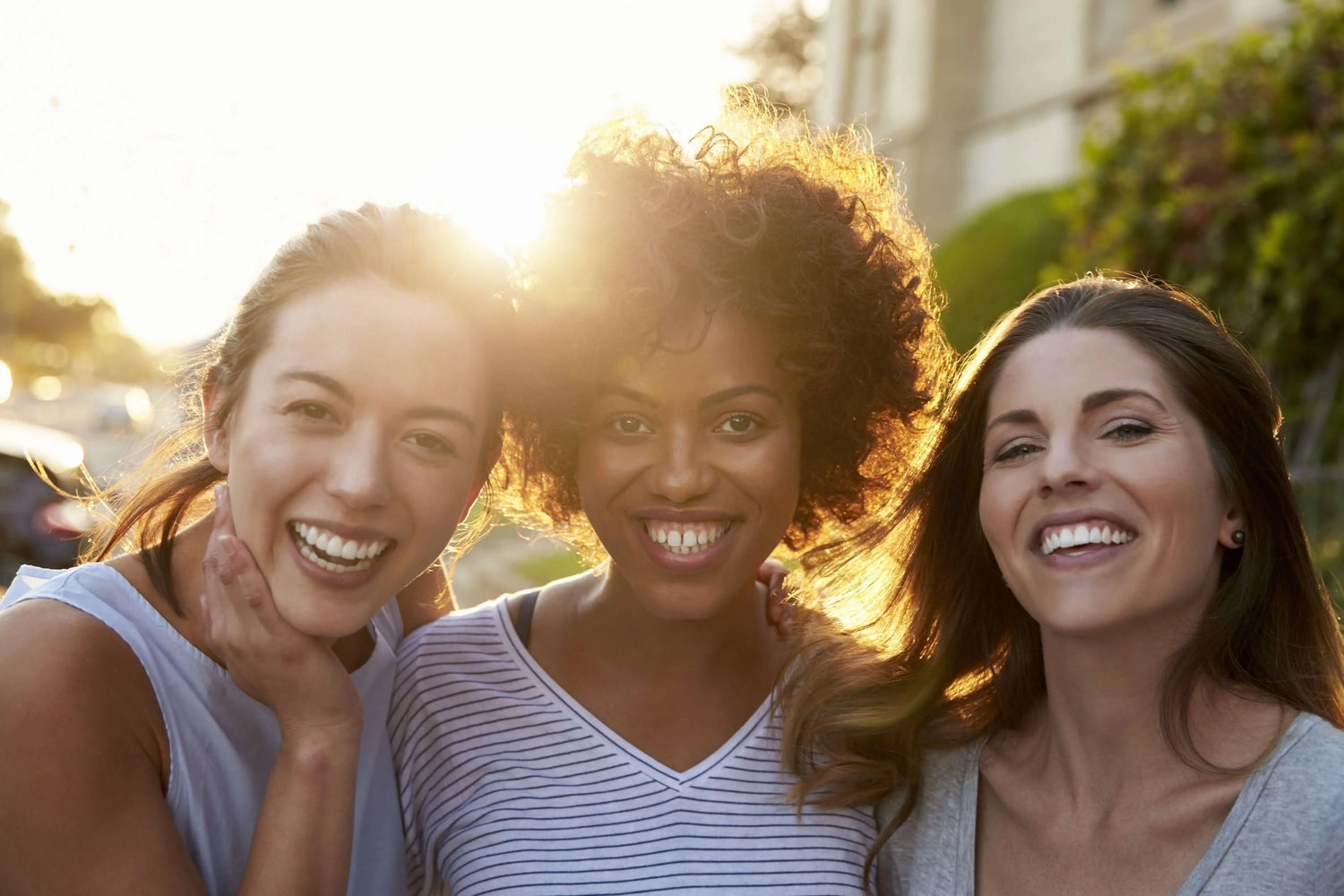 Três mulheres sorrindo ao ar livre sob a luz do sol, simbolizando saúde e bem-estar.