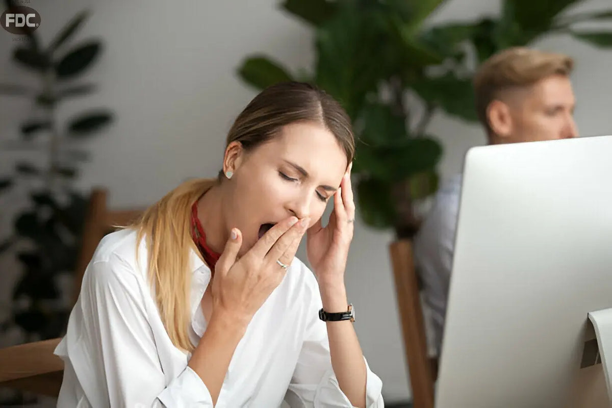 Texto alternativo:Mulher bocejando no trabalho, aparentando cansaço excessivo, com plantas ao fundo.