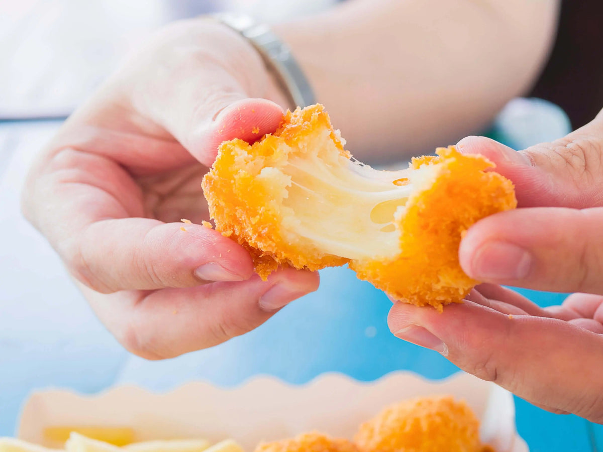 Texto alternativo:Pessoa segurando pedaço de queijo empanado derretido, alimento rico em gorduras.