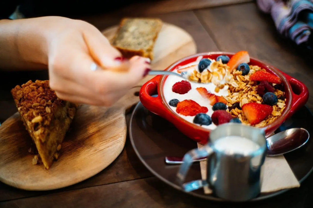 Texto alternativo:Tigela com iogurte, frutas vermelhas e granola, acompanhada de bolo saudável.