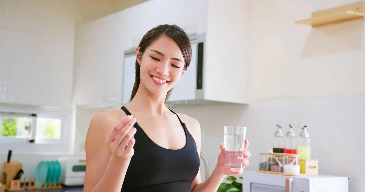 Texto alternativo:Mulher sorridente em roupa fitness segurando cápsula de Picolinato de Cromo e copo d'água na cozinha.