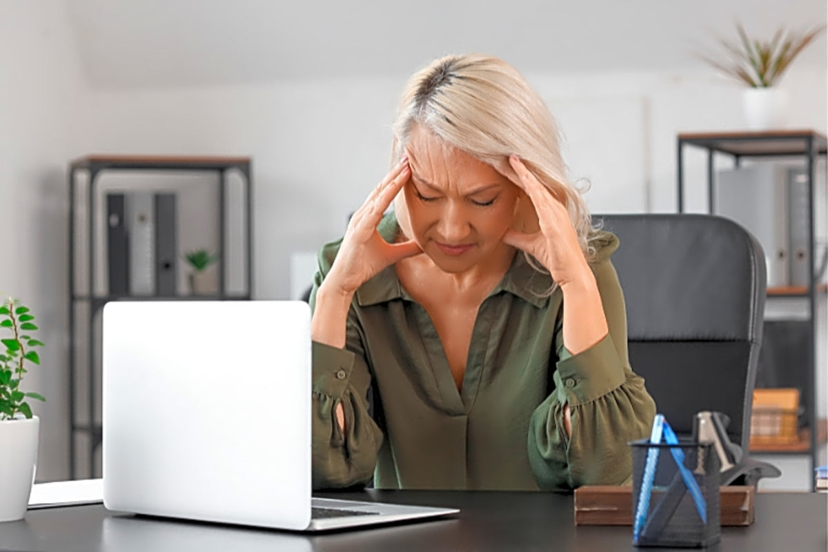 Mulher madura com expressão de desconforto, mãos na cabeça, sentada em frente ao computador.